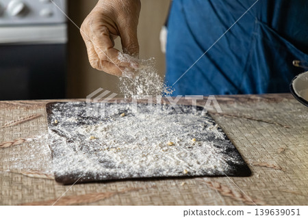 Elderly person hand sprinkling white flour onto a dark baking tray, preparing dough for traditional homemade bread or pastry, capturing authentic culinary craft and rustic kitchen atmosphere Elderly person hand sprinkling white flour onto a dark baking tray, preparing dough for traditional homemade bread or pastry, capturing authentic culinary craft and rustic kitchen atmosphere 139639051