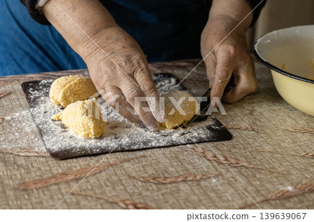 An elderly woman carefully prepares cottage cheese syrniki on a floured wooden board, shaping the dough for a traditional homemade breakfast that captures culinary craft and kitchen nostalgia 139639067