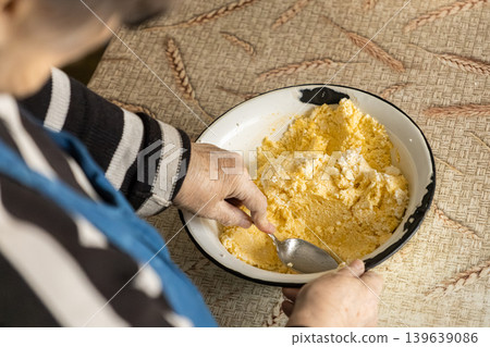 An elderly woman stirs a yellow cottage cheese mixture in an enamel bowl, preserving traditional home cooking methods and family heritage within an authentic, rustic kitchen culinary space An elderly woman stirs a yellow cottage cheese mixture in an enamel bowl, preserving traditional home cooking methods and family heritage within an authentic, rustic kitchen culinary space 139639086