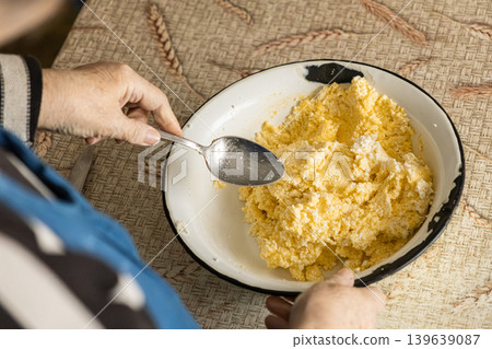 An elderly person stirs fresh cottage cheese and a raw egg in a metal bowl while preparing traditional dairy ingredients for authentic healthy family baking in a cozy rustic home kitchen An elderly person stirs fresh cottage cheese and a raw egg in a metal bowl while preparing traditional dairy ingredients for authentic healthy family baking in a cozy rustic home kitchen 139639087
