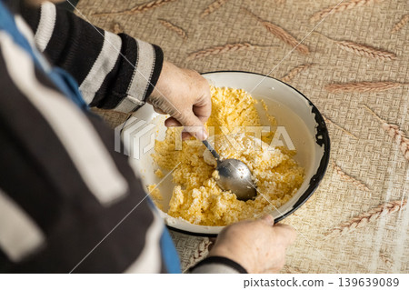 An elderly person stirs fresh homemade cottage cheese with a metal spoon in a white bowl, representing traditional culinary practices, organic food preparation, and authentic home cooking styles An elderly person stirs fresh homemade cottage cheese with a metal spoon in a white bowl, representing traditional culinary practices, organic food preparation, and authentic home cooking styles 139639089