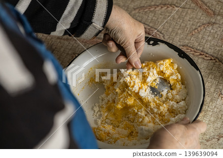 An elderly person stirs fresh cottage cheese with egg yolk in a bowl to prepare traditional homemade dough or filling for baking, representing authentic rustic culinary cooking traditions 139639094