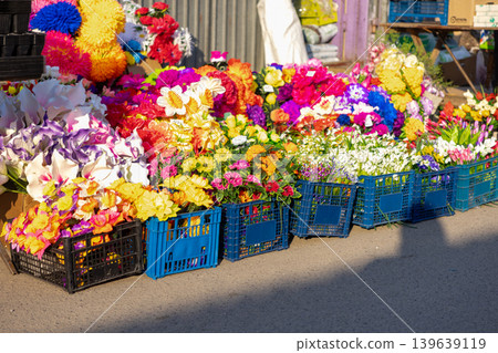 Colorful artificial flowers in plastic crates displayed for sale at an outdoor market stall, highlighting vibrant floral arrangements and wholesale retail themes in a casual urban shopping site Colorful artificial flowers in plastic crates displayed for sale at an outdoor market stall, highlighting vibrant floral arrangements and wholesale retail themes in a casual urban shopping site 139639119