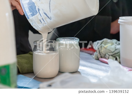 A person pours thick fresh sour cream from a large plastic bucket into glass jars for storage, showcasing artisanal food production, healthy dairy consumption and homemade kitchen craft style A person pours thick fresh sour cream from a large plastic bucket into glass jars for storage, showcasing artisanal food production, healthy dairy consumption and homemade kitchen craft style 139639152