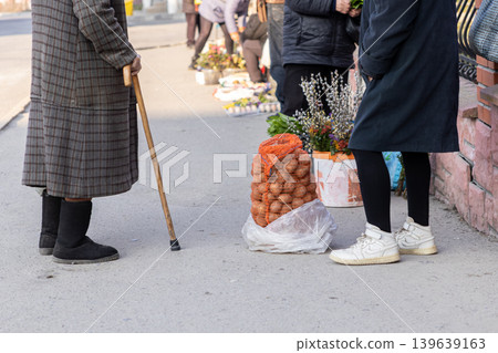 An elderly person with a cane and a customer stand by a bag of fresh potatoes at an outdoor street market to highlight local trade, small business economy, and rural life in a community An elderly person with a cane and a customer stand by a bag of fresh potatoes at an outdoor street market to highlight local trade, small business economy, and rural life in a community 139639163