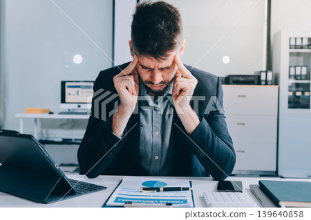 Tired worker. A tired businessman works in the evening. A man in office clothes is holding his temples. Problems in business. The businessman is trying to solve the problems that have arisen. 139640858