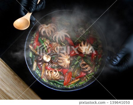 chef in black gloves cooking spicy stir-fried baby octopus with basil and chili in a hot pan with steam, high-angle view of delicious Asian seafood cuisine 139641050