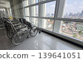 Empty wheelchairs lined up by a large window in a modern high-rise hospital corridor with a bright city view, symbolizing healthcare services and urban medical facilities 139641051