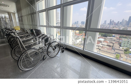Empty wheelchairs lined up by a large window in a modern high-rise hospital corridor with a bright city view, symbolizing healthcare services and urban medical facilities Empty wheelchairs lined up by a large window in a modern high-rise hospital corridor with a bright city view, symbolizing healthcare services and urban medical facilities 139641051