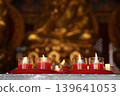 Red candles burning in glass cups on a prayer altar, with a blurred golden Buddha statue in the background. A symbol of faith, peace, and spiritual light in a Chinese temple 139641053
