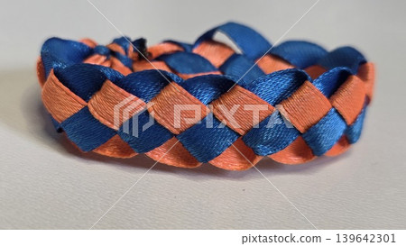 Handmade braided bracelet made of shiny blue and bright orange satin ribbons lying on plain white background, concept of creative kids craft and diy hobby Handmade braided bracelet made of shiny blue and bright orange satin ribbons lying on plain white background, concept of creative kids craft and diy hobby 139642301