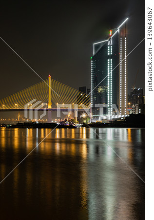 the striking, illuminated expanse of the Thotsamarachan Bridge (Rama IX Bridge) spans the Chao Phraya River alongside a sleek, modern skyscraper, both casting vibrant golden reflections across the the striking, illuminated expanse of the Thotsamarachan Bridge (Rama IX Bridge) spans the Chao Phraya River alongside a sleek, modern skyscraper, both casting vibrant golden reflections across the 139643067