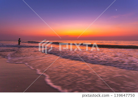 Serene beach sunset with a lone figure and gentle waves lapping the shore Serene beach sunset with a lone figure and gentle waves lapping the shore 139647750