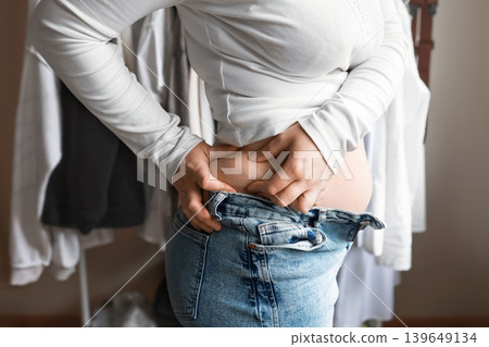 Side view of an overweight woman pinching extra fat on her waist while wearing tight, unbuttoned jeans, showcasing weight gain and clothing problems. Side view of an overweight woman pinching extra fat on her waist while wearing tight, unbuttoned jeans, showcasing weight gain and clothing problems. 139649134
