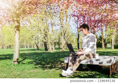 A man sitting on a park bench working on his laptop under blooming pink cherry blossom trees, enjoying remote work in spring. A man sitting on a park bench working on his laptop under blooming pink cherry blossom trees, enjoying remote work in spring. 139649136