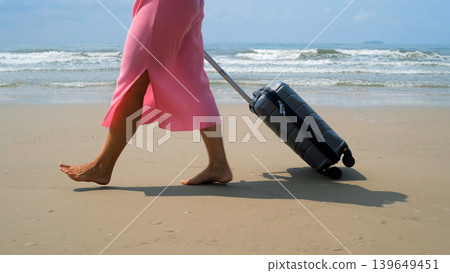Young barefoot woman in pink dress rolling a grey suitcase along sandy beach with blue sea and waves in background. Summer travel concept 139649451