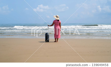 Rear view of young woman in pink dress and straw hat standing with suitcase at the water edge on sandy beach facing the sea. Arrival vacation concept Rear view of young woman in pink dress and straw hat standing with suitcase at the water edge on sandy beach facing the sea. Arrival vacation concept 139649458