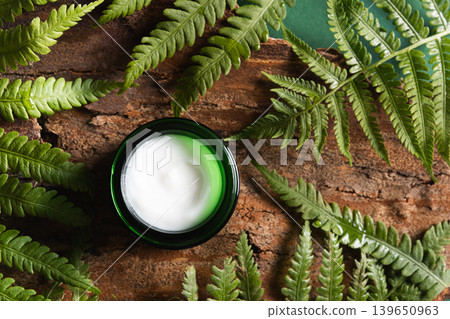Cosmetic face cream products in open green glass jar with natural ingredients decorated fresh fern leaves and wood bark. Cosmetic face cream products in open green glass jar with natural ingredients decorated fresh fern leaves and wood bark. 139650963