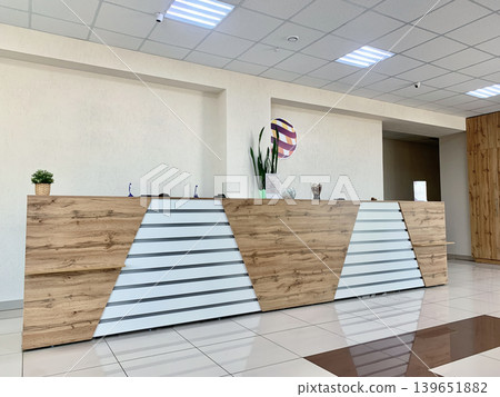 Modern reception desk showcases a wooden finish with geometric patterns in a bright office space, complemented by indoor plants and decorative items 139651882