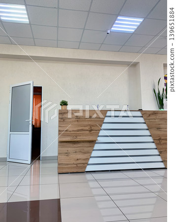 Modern reception area features a wooden front desk, an open door leading to another room, and a potted plant on the counter Modern reception area features a wooden front desk, an open door leading to another room, and a potted plant on the counter 139651884