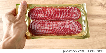 Hand holding a pack of sliced salami on a wooden table mid day Hand holding a pack of sliced salami on a wooden table mid day 139652890