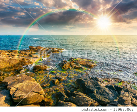 sunset over sea coast with stones and boulders. view of beach with rock in seaweed. dynamic cloudscape on the sky with rainbow above horizon afternoon. summer vacations background concept sunset over sea coast with stones and boulders. view of beach with rock in seaweed. dynamic cloudscape on the sky with rainbow above horizon afternoon. summer vacations background concept 139653062