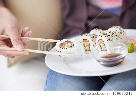 Hands, sushi plate and chopsticks in closeup, home or eating for wellness, diet or meal with soy sauce. Hungry person, fish and dish at lunch, nutrition or snack for takeaway food, house or apartment Hands, sushi plate and chopsticks in closeup, home or eating for wellness, diet or meal with soy sauce. Hungry person, fish and dish at lunch, nutrition or snack for takeaway food, house or apartment 139653212