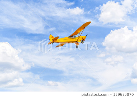 A yellow single-engine ultralight aircraft flying in a blue sky with white clouds. A yellow single-engine ultralight aircraft flying in a blue sky with white clouds. 139653870