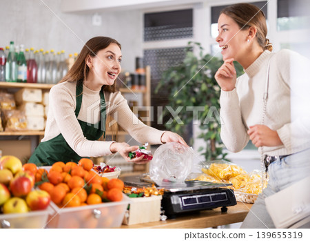 Woman sells and puts candies in bag for young woman, puts them on scale and weighs fruits Woman sells and puts candies in bag for young woman, puts them on scale and weighs fruits 139655319