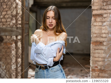 Woman in shirt pulled down from shoulders standing near wire fencing in abandoned house Woman in shirt pulled down from shoulders standing near wire fencing in abandoned house 139655320