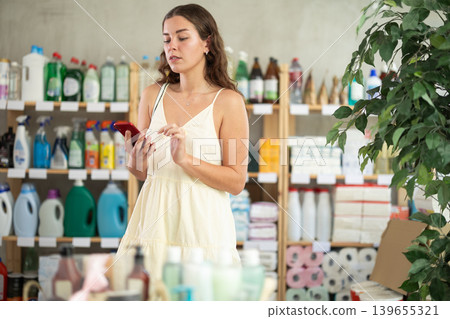 Female shopper searches for necessary goods on the shelves of a supermarket, checking the list on smartphone Female shopper searches for necessary goods on the shelves of a supermarket, checking the list on smartphone 139655321