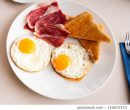 Fried eggs with slices of ham and wheat bread toasts Fried eggs with slices of ham and wheat bread toasts 139655553