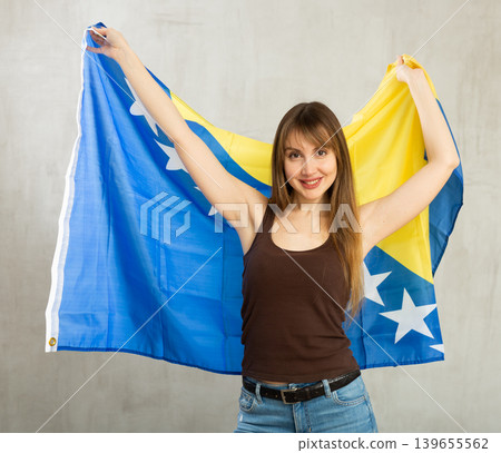 Happy cheerful young woman waving national flag of Bosnia and Herzegovina while looking at camera at gray wall background Happy cheerful young woman waving national flag of Bosnia and Herzegovina while looking at camera at gray wall background 139655562