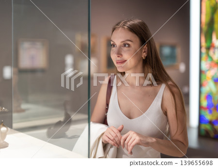 Young female visitor stands near glass display case and examines valuable exhibit Young female visitor stands near glass display case and examines valuable exhibit 139655698