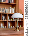 A stylish table lamp sits on a wooden surface beside a bookshelf filled with neatly arranged books, creating a cozy reading atmosphere in natural light. 139656904