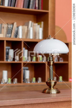 A stylish table lamp sits on a wooden surface beside a bookshelf filled with neatly arranged books, creating a cozy reading atmosphere in natural light. 139656904