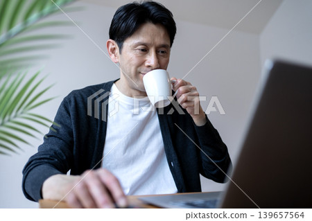 A middle-aged man using his laptop with a cup of coffee in hand during his morning routine. A middle-aged man using his laptop with a cup of coffee in hand during his morning routine. 139657564