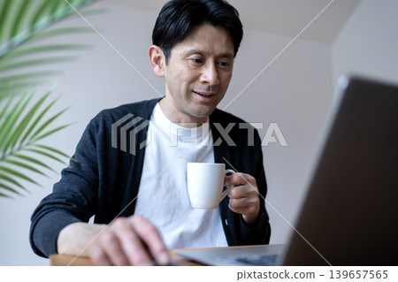 一位中年男子在晨間例行活動中，一邊手持咖啡，一邊使用筆記型電腦。 139657565