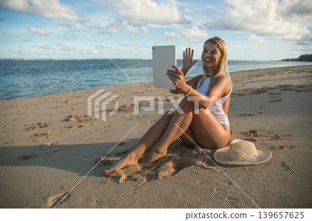Woman making a video call while sitting on the sand Woman making a video call while sitting on the sand 139657625