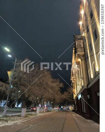 A stunning evening view featuring a brightly lit building facade against a dark sky, with trees lining the street. Perfect for urban planning, travel, or architectural design. 139658397