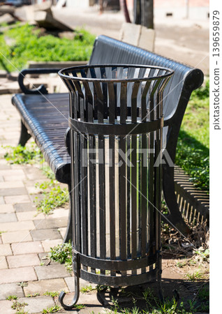 Outdoor metal trash bin beside park bench on sunny paved walkway. Outdoor metal trash bin beside park bench on sunny paved walkway. 139659879