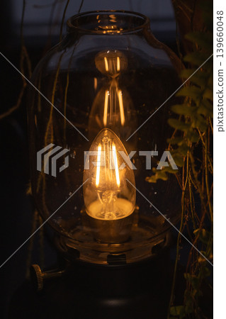 A warm vintage filament lamp glows inside a glass lantern, casting cozy amber light A warm vintage filament lamp glows inside a glass lantern, casting cozy amber light 139660048