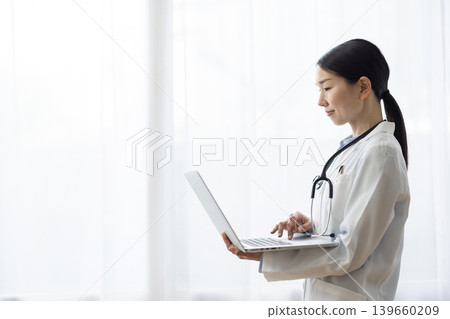 A Japanese female doctor in a white coat is operating a laptop computer by a bright window. A Japanese female doctor in a white coat is operating a laptop computer by a bright window. 139660209