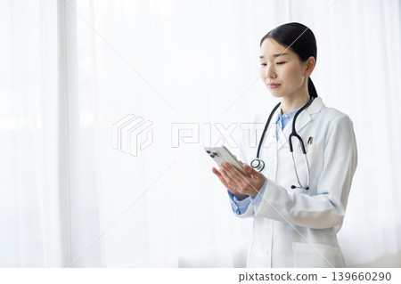 一位身穿白大褂的日本女醫生正在明亮的窗邊操作智慧型手機。 139660290