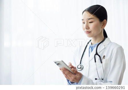 A Japanese female doctor in a white coat is operating her smartphone by a bright window. A Japanese female doctor in a white coat is operating her smartphone by a bright window. 139660291