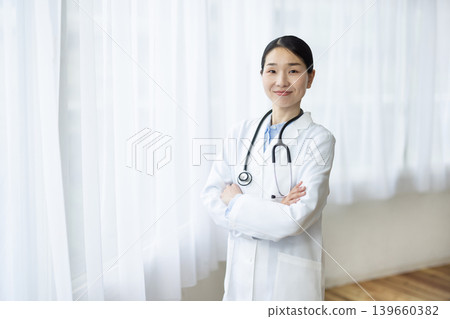 一位身穿白大褂的日本女醫生站在明亮的窗前，雙手交叉，面帶微笑。 139660382