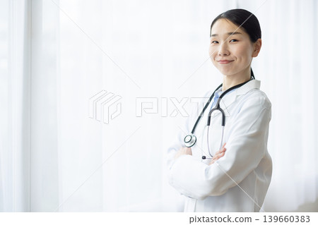 A Japanese female doctor in a white coat smiles with her arms crossed by a bright window. A Japanese female doctor in a white coat smiles with her arms crossed by a bright window. 139660383