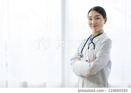 A Japanese female doctor in a white coat smiles with her arms crossed by a bright window. A Japanese female doctor in a white coat smiles with her arms crossed by a bright window. 139660385