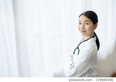 A Japanese female doctor in a white coat smiles with her arms crossed by a bright window. A Japanese female doctor in a white coat smiles with her arms crossed by a bright window. 139660386