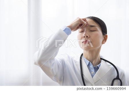A Japanese female doctor in a white coat touches her temple and makes a pained expression. A Japanese female doctor in a white coat touches her temple and makes a pained expression. 139660994